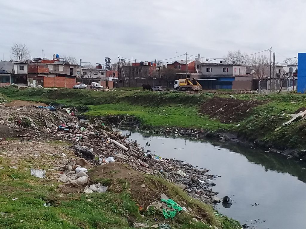 se observa el canal José León Suárez localizado en el Barrio Carcova, popularmente conocido como el zanjón. la imagen fue tomada en uno de los puntos de muestreo de aguas y sedimentos. se observa un ambiente degradado con basurales irregurales en las orillas del canal, en la orilla de enfrente se pueden ver los sedimentos que se depositan en el recodo que se forma con el correr del tiempo, sobre los sedimentos crece vegetación bastante verde, alli crecen unos pastizales en dónde habitan algunas aves con patas alargadas, típicas de humedal. esta imagen nos permite percibir como la naturaleza aún persiste en cuidar la vida que resiste a pasar de la contaminación